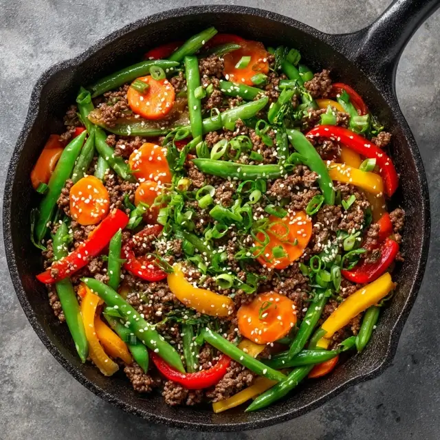A skillet filled with ground beef stir fry, featuring colorful bell peppers, green beans, and carrots, garnished with sesame seeds and green onions.